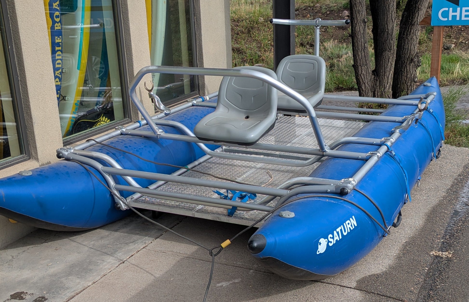 A 16' Cataraft Saturn by 4Corners Riversports, featuring a metal frame and two seats, is parked on a sidewalk beside a building with large windows.