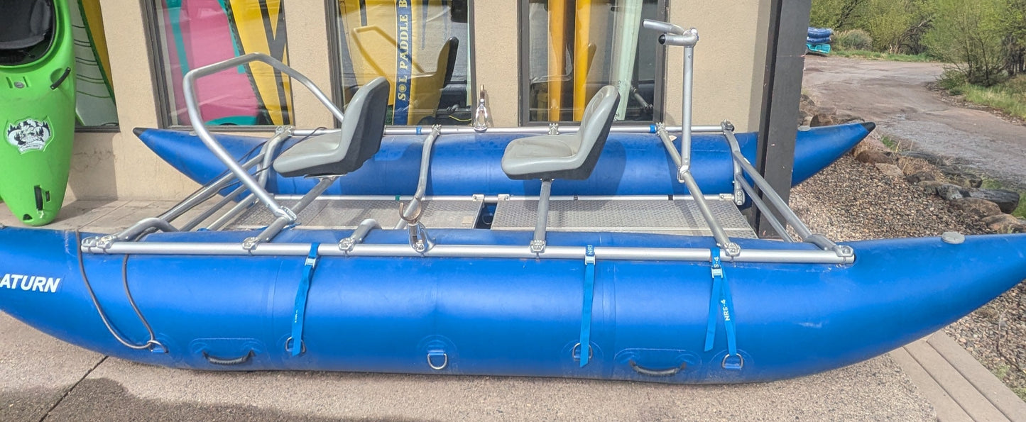 A 16' Cataraft Saturn by 4Corners Riversports with two seats is parked outside a building, where colorful kayaks and paddles can be seen through the windows.