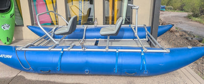 A 16' Cataraft Saturn by 4Corners Riversports with two seats is parked outside a building, where colorful kayaks and paddles can be seen through the windows.