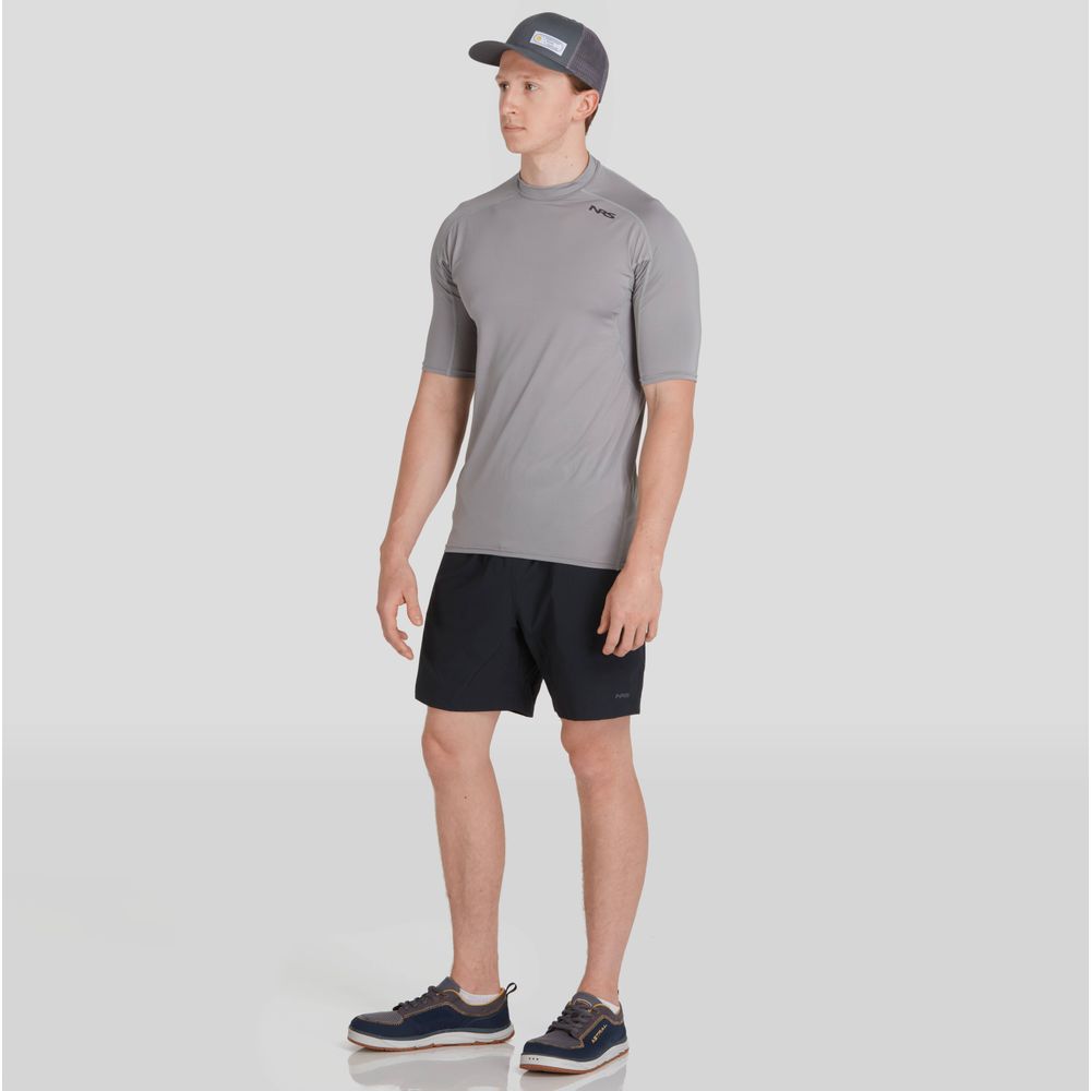 A man in an NRS Short-Sleeve Rashguard and gray cap, paired with black shorts and sneakers, stands against a plain background.