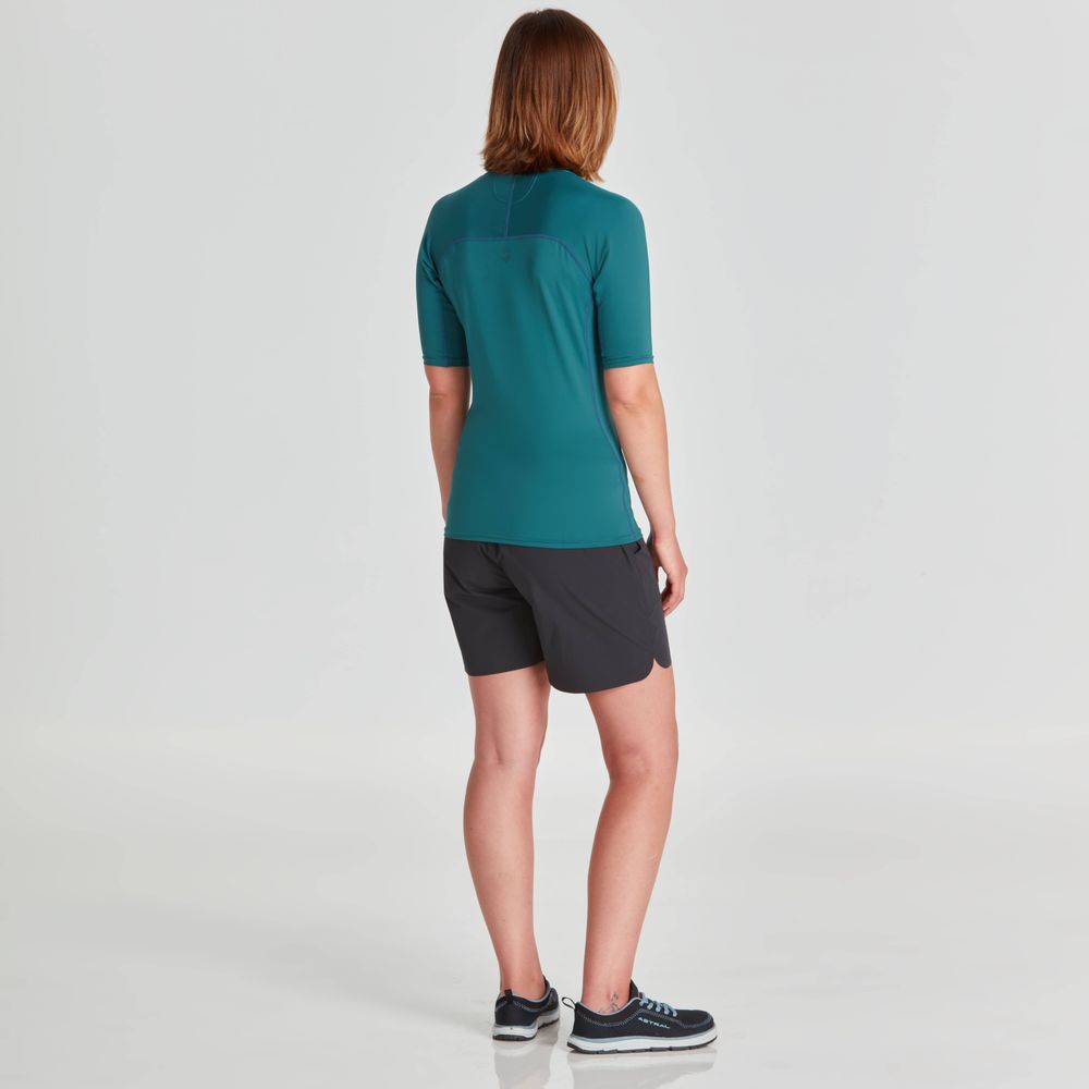 A person with shoulder-length hair faces away from the camera, sporting NRS's Short Sleeve Rashguard - Women's in teal and black shorts. Their black sneakers stand out against the plain white backdrop, emphasizing the outfit's stylish design and sun protection features.