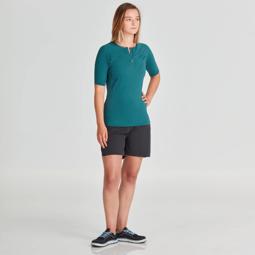 A person is wearing an NRS Short Sleeve Rashguard in teal paired with black shorts and black sneakers against a plain white background.