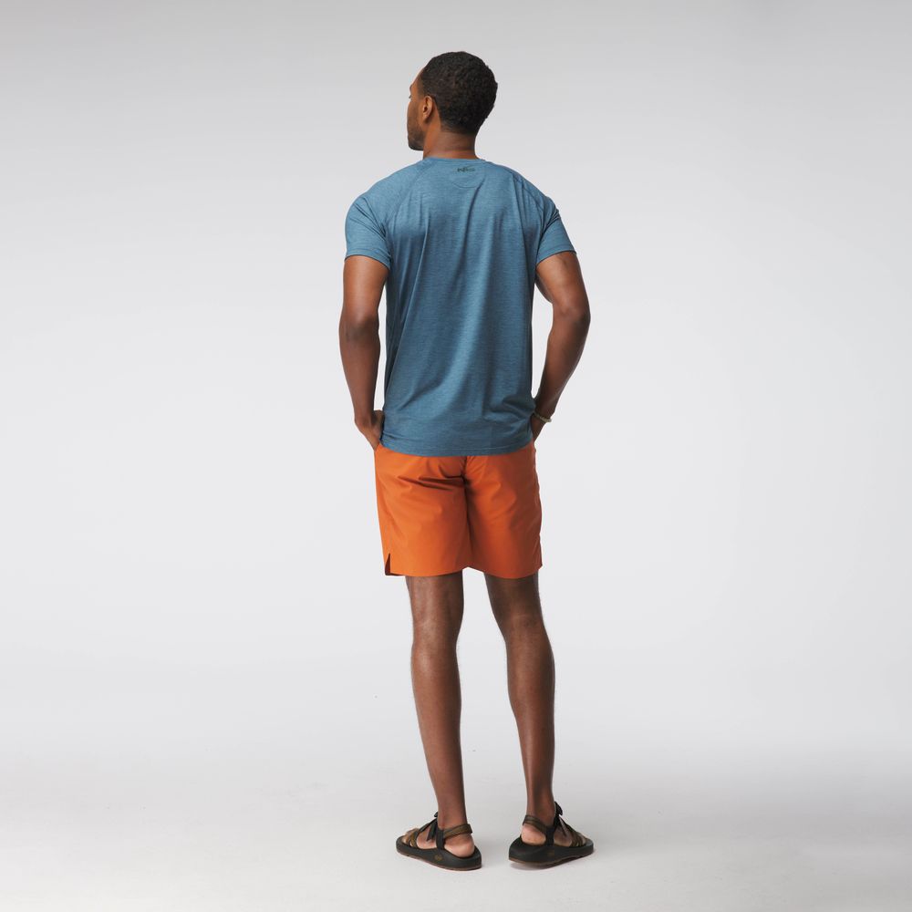 A man in an NRS Silkweight Short-Sleeve Shirt and orange shorts stands with his back to the camera, hands in pockets, against a plain backdrop. The moisture-wicking fabric provides sun protection for warm days.
