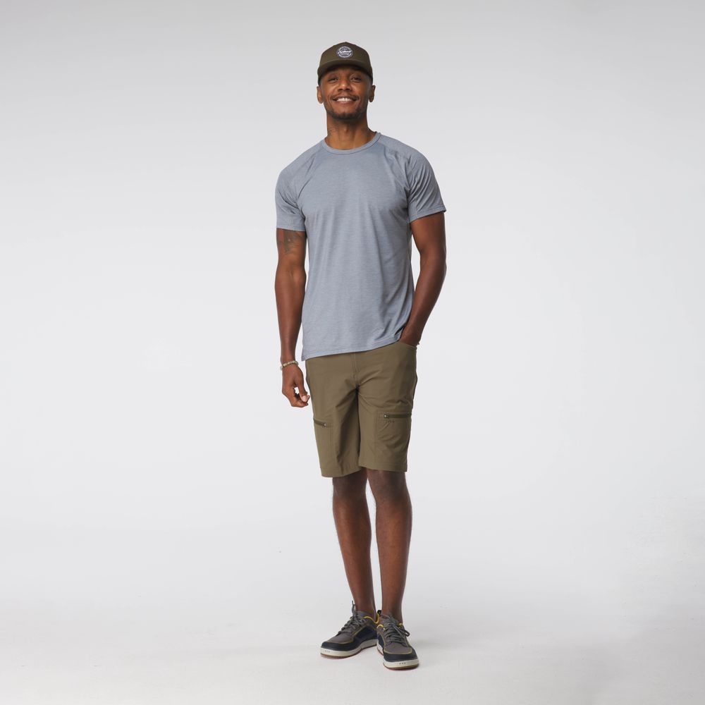 A man wearing an NRS Silkweight Short-Sleeve Shirt, olive cargo shorts, and a cap smiles contentedly, relishing the comfort of the moisture-wicking fabric against a white background.