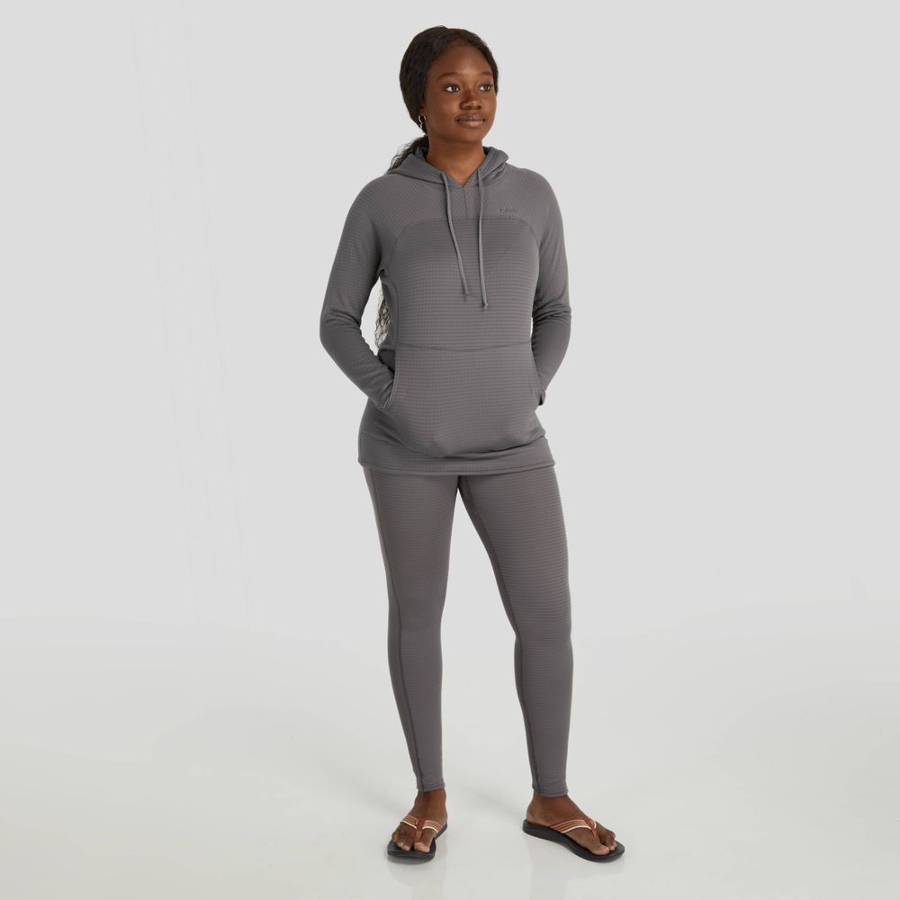 A woman stands on a light background in a gray NRS Lightweight Hoodie, featuring moisture-wicking fabric and UPF 50+ protection, with hands in pockets. She pairs it with leggings and sandals.