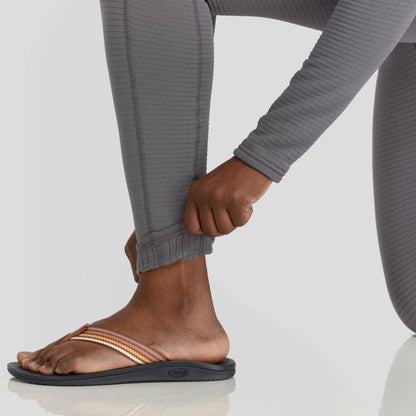 A woman in NRS Lightweight Pants with moisture-wicking fabric adjusts the ankle hem while wearing multicolored sandals on a gray backdrop.