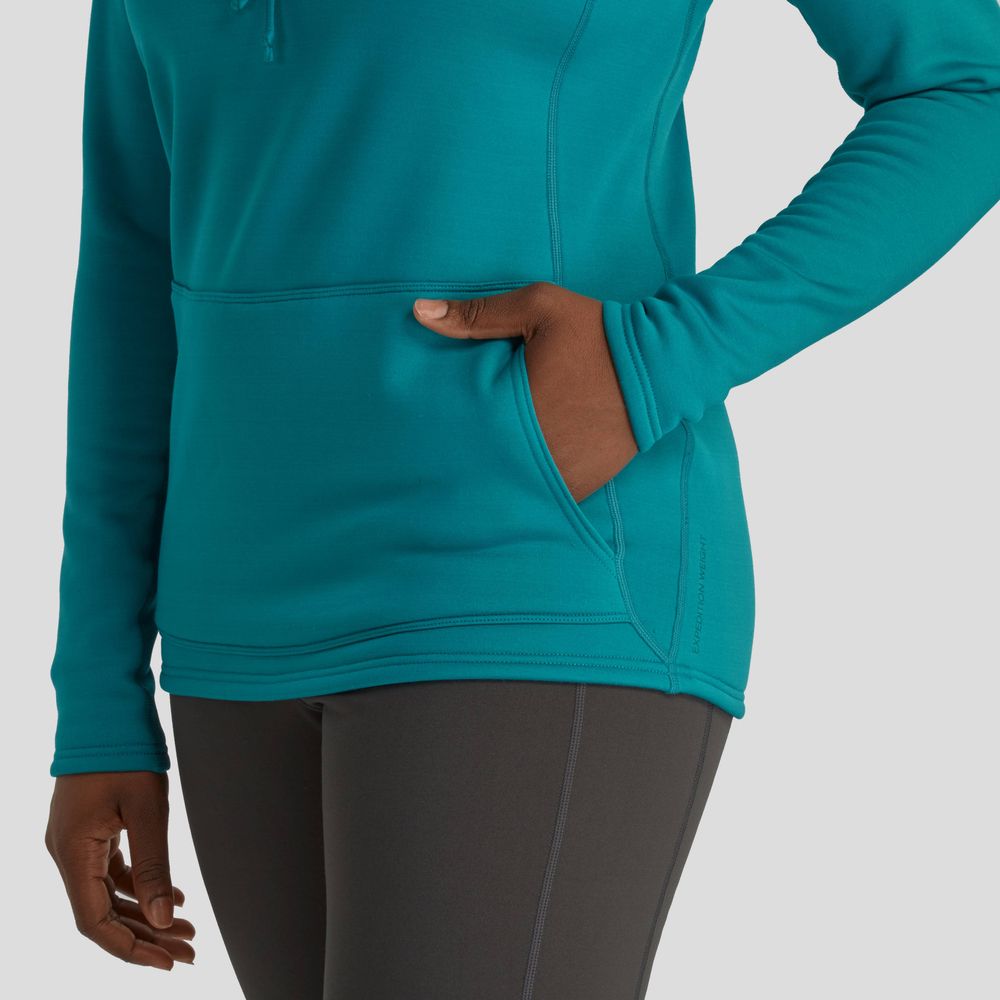 A person wears a teal NRS Expedition Hoodie for women, featuring long sleeves, a kangaroo pocket, and paired with dark grey leggings. This high-stretch fleece offers breathable insulation against any plain backdrop.