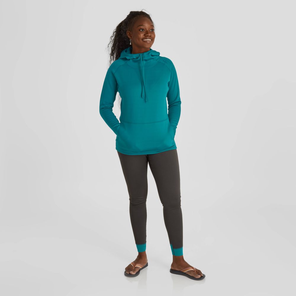 A woman stands smiling, wearing a NRS Expedition Hoodie - Women's in teal with breathable insulation, dark leggings, and sandals against a plain background.