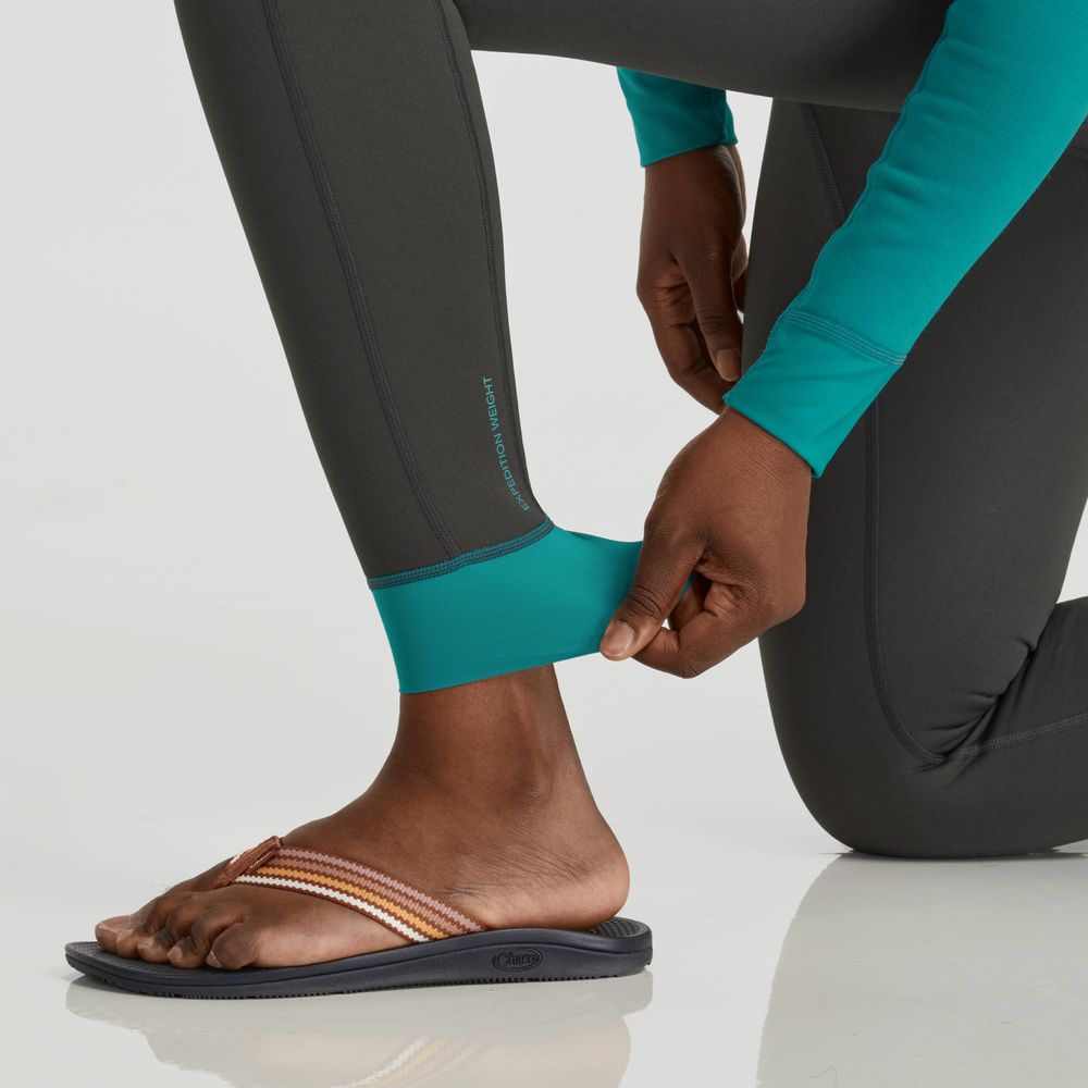 A woman adjusts the cuff of her teal and gray NRS Expedition Weight Pants while kneeling, paired with multicolored flip-flops.