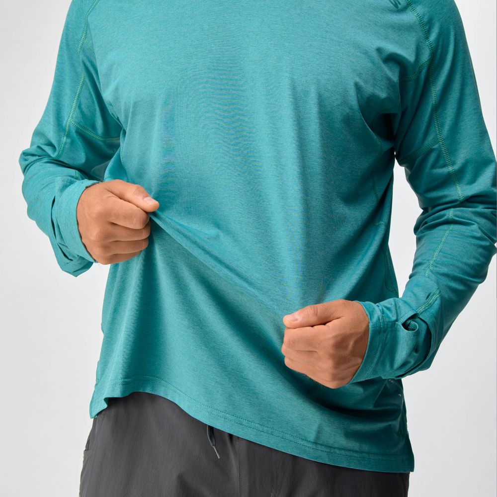 A person wearing the NRS Silkweight Hoodie - Men's in teal with gray pants, holding and slightly stretching the moisture-wicking long-sleeve shirt.
