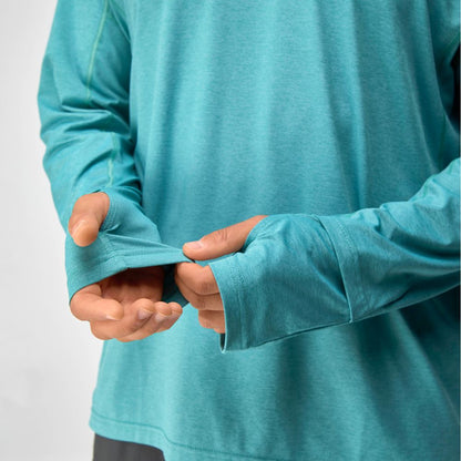 A man wears the NRS Silkweight Hoodie - Men's in teal, pulling on the sleeve fabric with both hands. This moisture-wicking hoodie features thumbholes and offers sun protection and comfort for outdoor activities.