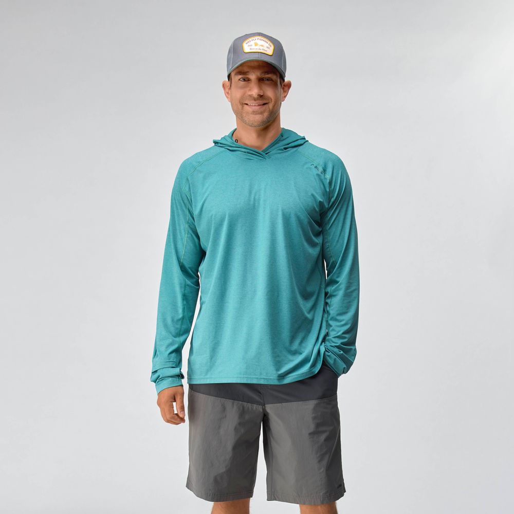 A man wearing the NRS Silkweight Hoodie - Men's in teal, paired with gray shorts and a gray cap, stands against a plain light background, smiling at the camera.
