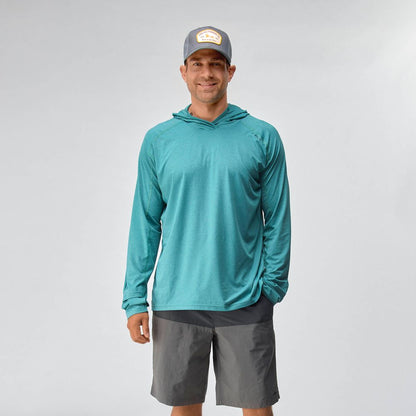 A man wearing the NRS Silkweight Hoodie - Men's in teal, paired with gray shorts and a gray cap, stands against a plain light background, smiling at the camera.