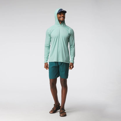 A man smiles in a light blue NRS Silkweight Hoodie - Men's, which provides sun protection, paired with teal shorts, sandals, and a blue cap against a plain light background.
