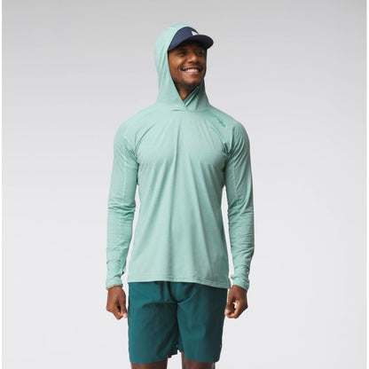 A man smiles against a plain light background, wearing the NRS Silkweight Hoodie - Men's in light green for sun protection, paired with dark green shorts and a navy cap.