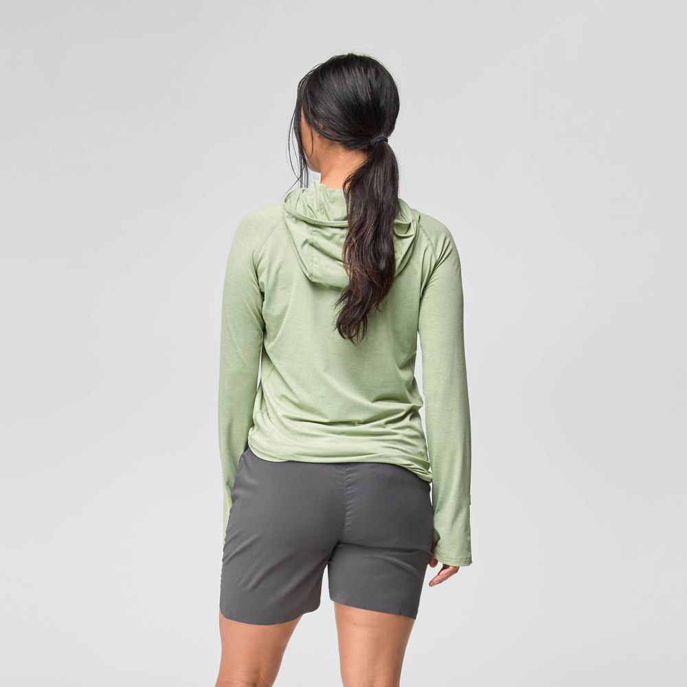 A woman faces away, wearing the NRS Silkweight Hoodie - Women's—a light green sun-protective baselayer—paired with dark gray shorts against a plain, light background.