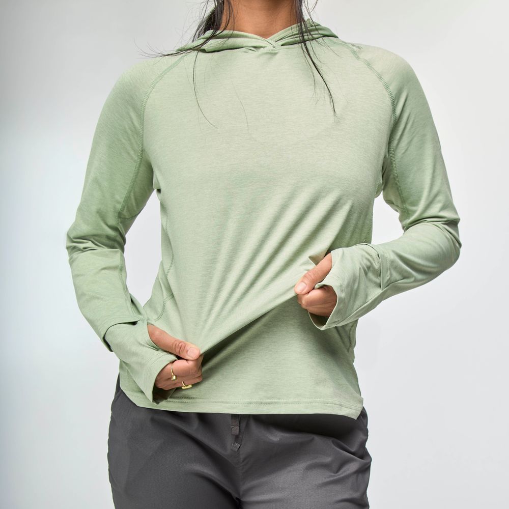 A person wears the NRS Silkweight Hoodie - Women's in light green with dark pants, holding the hem—ideal as a sun-protective base layer against a plain background.