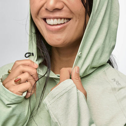 A smiling person in the NRS Silkweight Hoodie - Women's adjusts the hood, showing sun protection and comfort in this light green women's base layer; only the lower half of their face is visible.