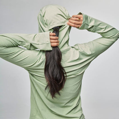 A woman with long dark hair, seen from behind, pulls the hood of her light green NRS Silkweight Hoodie over her head for sun protection.