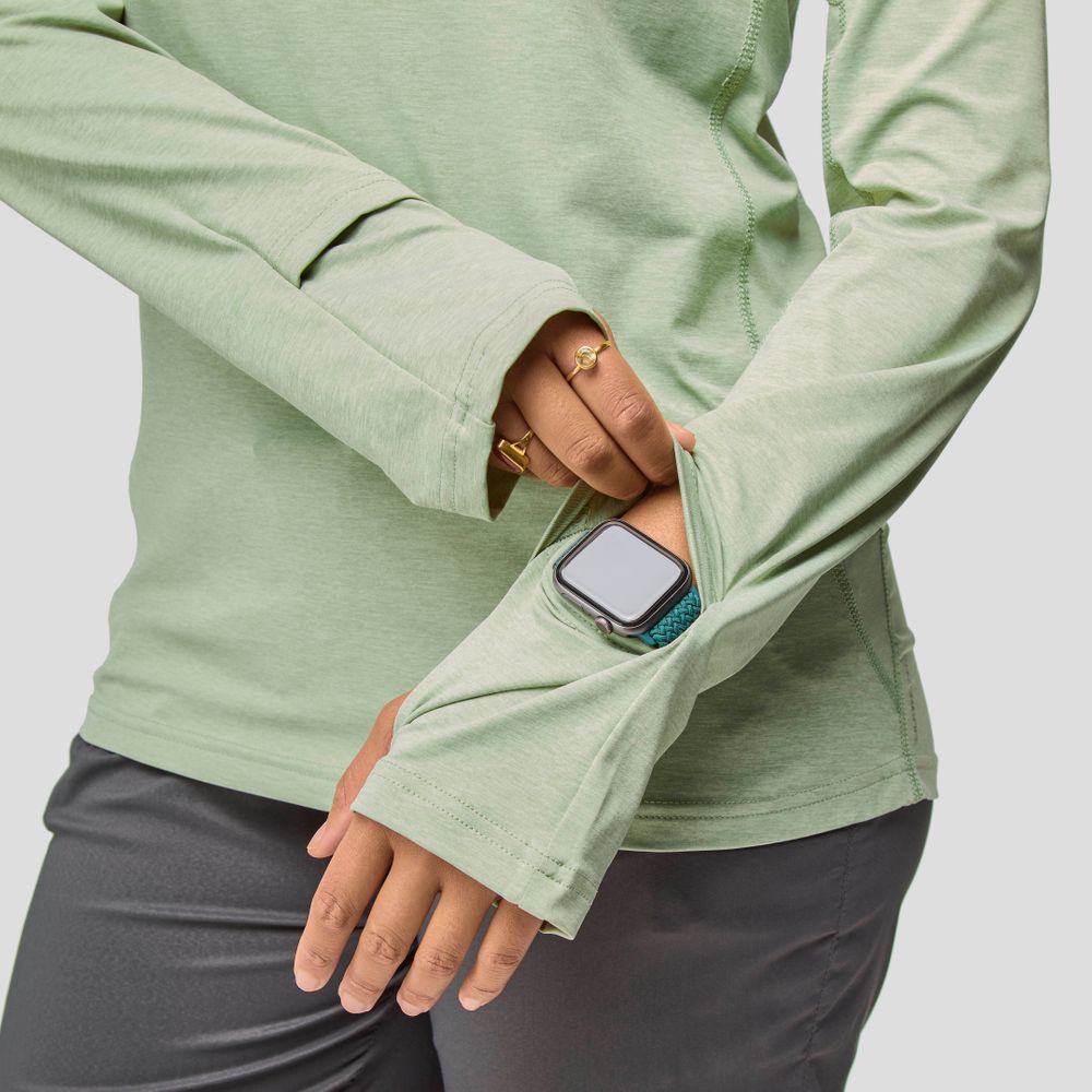 A woman in an NRS Silkweight Hoodie shows off a smartwatch through the thumbhole cuff, pairing light green sun protection with dark pants for a stylish look.