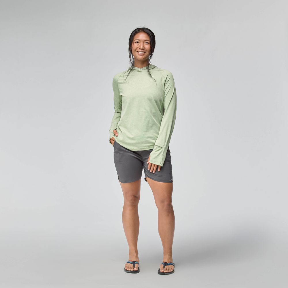 A person stands facing forward, smiling, wearing the NRS Silkweight Hoodie - Women's for sun protection with dark shorts and flip-flops against a plain light background.