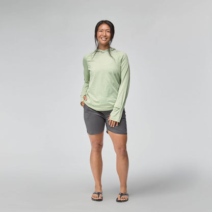 A person stands facing forward, smiling, wearing the NRS Silkweight Hoodie - Women's for sun protection with dark shorts and flip-flops against a plain light background.