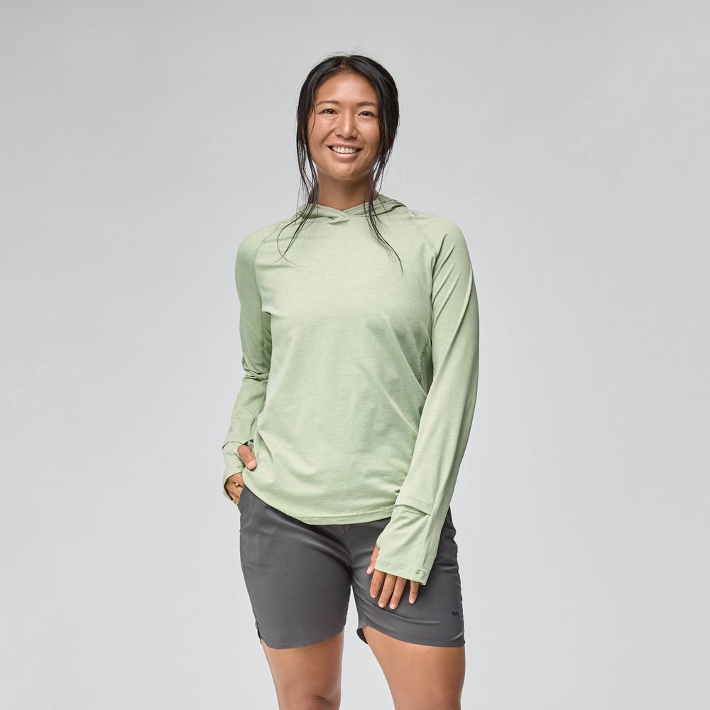 A woman smiles against a plain background, wearing a light green NRS Silkweight Hoodie - Women's for sun protection and gray shorts, with her left hand in her pocket.