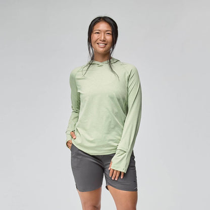 A woman smiles against a plain background, wearing a light green NRS Silkweight Hoodie - Women's for sun protection and gray shorts, with her left hand in her pocket.