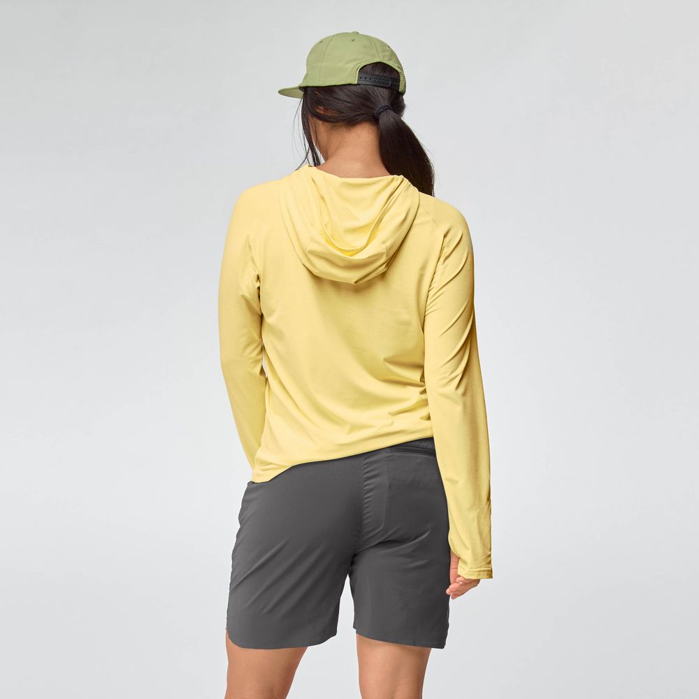 A woman stands with her back to the camera, wearing the NRS Silkweight Hoodie - Women's in yellow for sun protection, a light green cap, and dark gray shorts against a plain light background.