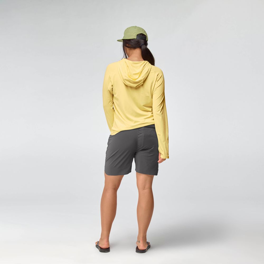 A person faces away, wearing the NRS Silkweight Hoodie - Women's in light yellow for sun protection, paired with gray shorts, a green cap, and flip-flops against a plain light background.