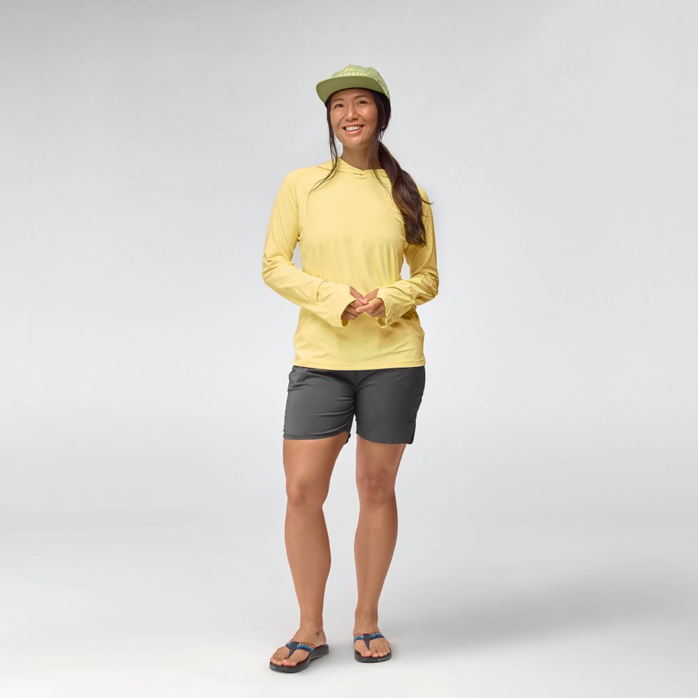 A person smiles against a plain light background, wearing an NRS Silkweight Hoodie - Women's in yellow, gray shorts, flip-flops, and a green cap.