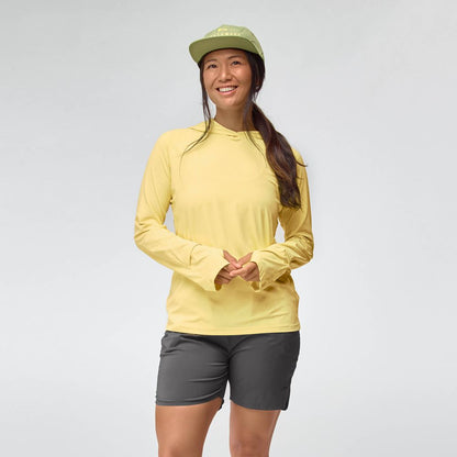 A woman wearing the NRS Silkweight Hoodie - Women's in yellow, gray shorts, and a green cap stands smiling against a plain light background.