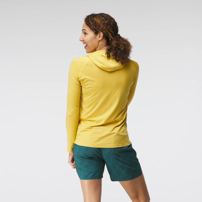 A woman wearing a yellow NRS Silkweight Hoodie and green patterned shorts stands partially turned, smiling. The technical fabric provides excellent sun protection for sunny outdoor days.