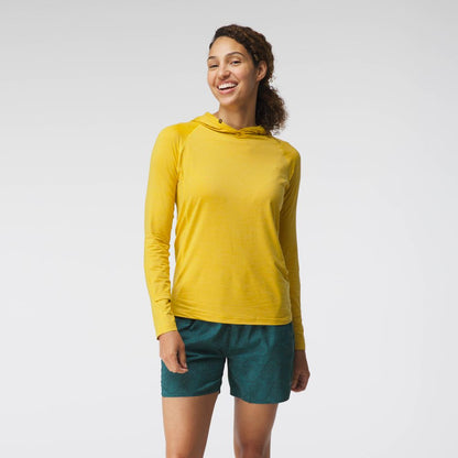 A woman smiles in an NRS Silkweight Hoodie - Women's for sun protection, paired with green patterned shorts, against a plain white background.