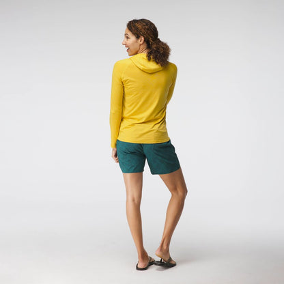 A woman faces away in an NRS Silkweight Hoodie for sun protection, paired with green shorts and flip-flops, set against a plain white background.