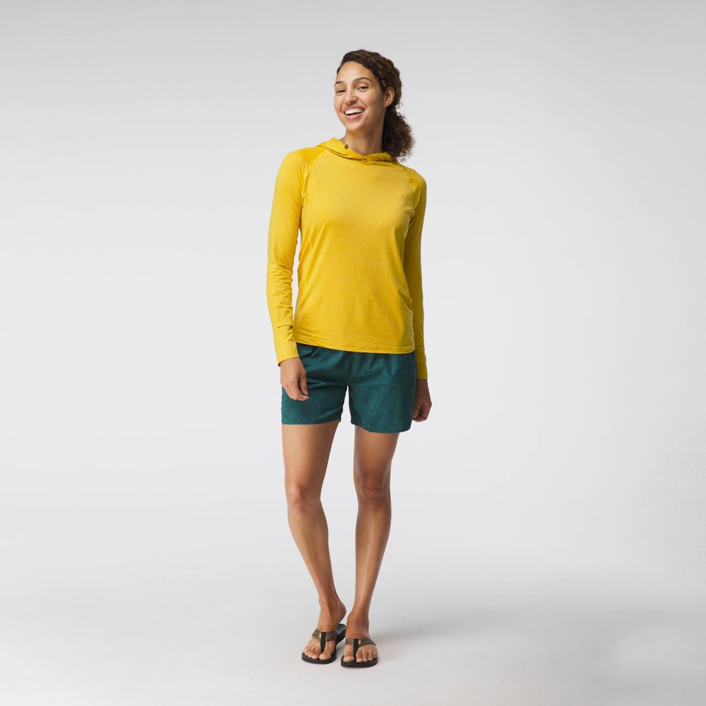 A woman stands against a plain background wearing the NRS Silkweight Hoodie—her sun-protective top is the perfect base layer for outdoor adventures, paired with teal shorts and sandals.