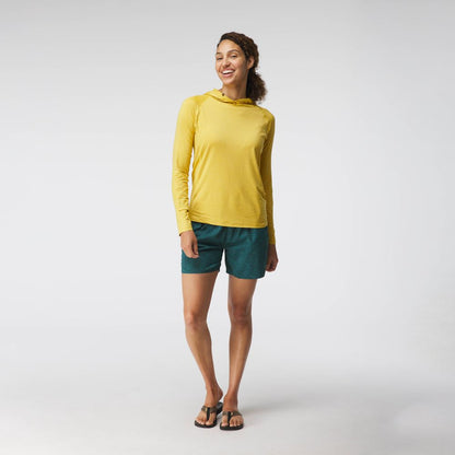 A person smiles, wearing a yellow NRS Silkweight Hoodie for sun protection, teal shorts, and black sandals against a light gray background.