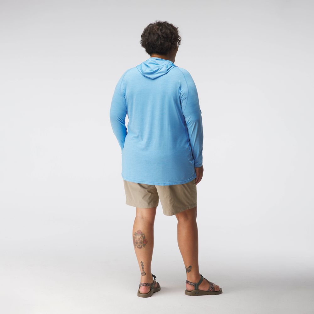 A woman stands with her back to the camera in a light blue NRS Silkweight Hoodie - Women's for sun protection, khaki shorts, and sandals. Tattoos are visible on both legs. The background is plain white.