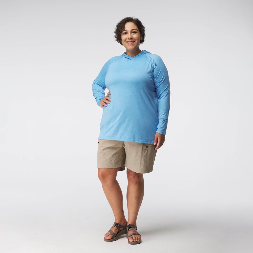A woman faces forward against a white background, wearing the NRS Silkweight Hoodie - Women's in light blue for sun protection, paired with tan shorts and sandals.
