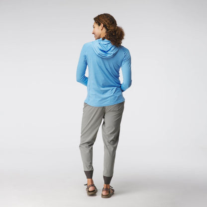 A person with curly hair stands against a light background, wearing gray pants, sandals, and a blue NRS Silkweight Hoodie - Women's, designed with technical fabrics for comfort and sun protection.