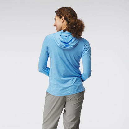 Facing away, a person in a blue NRS Silkweight Hoodie - Women's, made from technical fabrics, stands with hands on hips in gray pants against a plain backdrop, ready for sun protection.