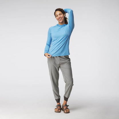 A woman wearing the NRS Silkweight Hoodie in light blue, which offers sun protection, along with gray pants and sandals, poses with one hand on her head against a plain background.