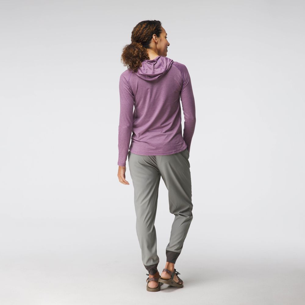 A woman stands facing away, wearing the NRS Silkweight Hoodie - Women's in light purple, with gray jogger pants and sandals, set against a plain light background.