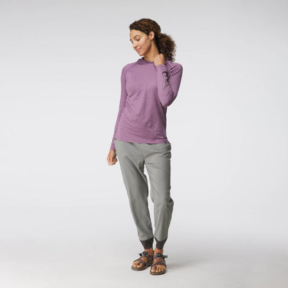 A woman stands in a relaxed pose wearing the NRS Silkweight Hoodie - Women's, paired with gray pants and sandals, against a plain white background.