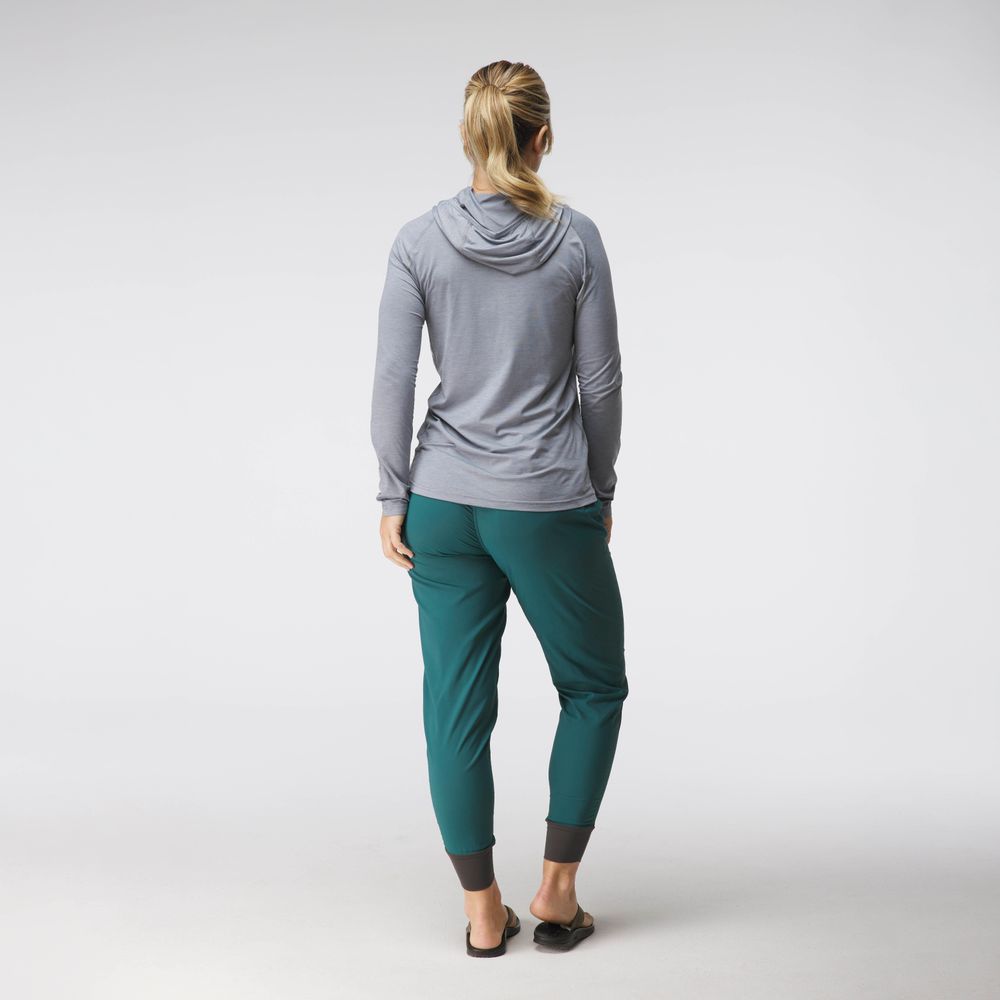 A woman faces away, wearing the NRS Silkweight Hoodie - Women's in light gray for sun protection, teal jogger pants, and black slip-on shoes against a plain light background.