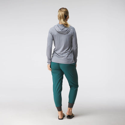 A woman faces away, wearing the NRS Silkweight Hoodie - Women's in light gray for sun protection, teal jogger pants, and black slip-on shoes against a plain light background.