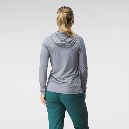 A woman stands with her back to the camera, wearing an NRS gray Silkweight Hoodie and teal technical-fabric pants against a plain background.