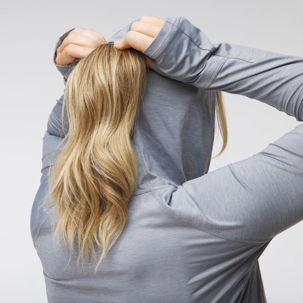 A woman with long blonde hair, seen from behind, pulls up the hood of an NRS Silkweight Hoodie - Women's in light gray for sun protection.