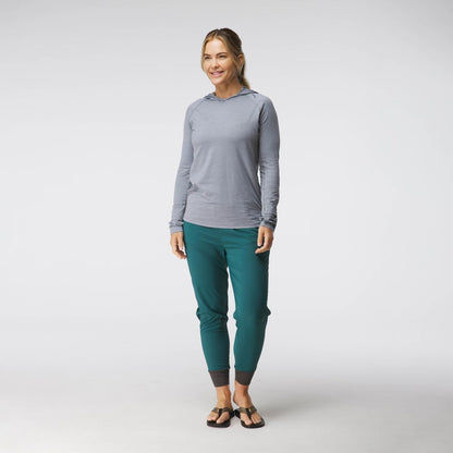 A woman in a casual gray NRS Silkweight Hoodie and teal pants smiles in a studio setting, wearing black sandals. Her light brown hair is pulled back, showcasing the comfort and sun protection of technical fabrics.