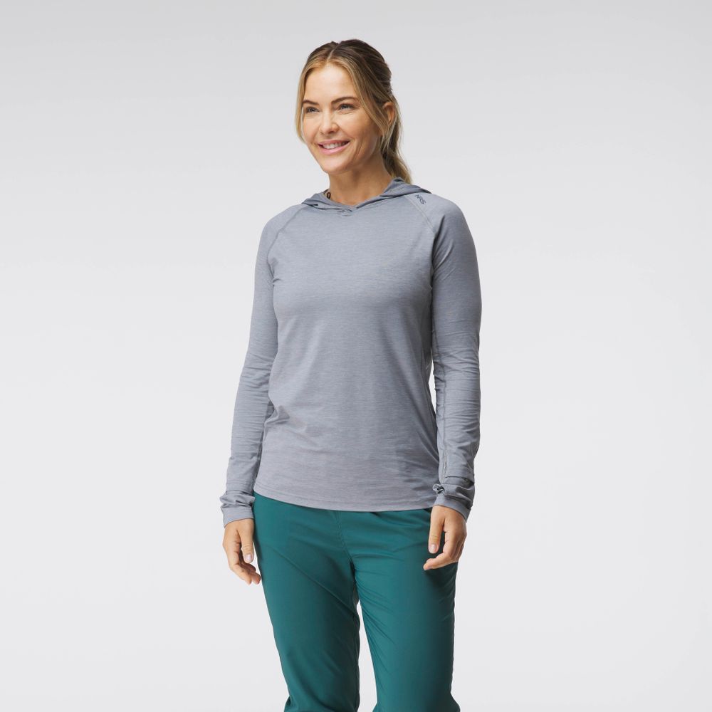 A woman stands smiling in the NRS Silkweight Hoodie - Women's in light gray and teal pants, ideal as a sun-protective base layer, set against a plain white background.